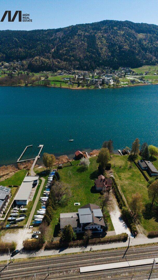Vertikale Luftaufnahme eines Seegrundstücks mit Häusern, Bahngleisen und dem weiten See im Vordergrund.