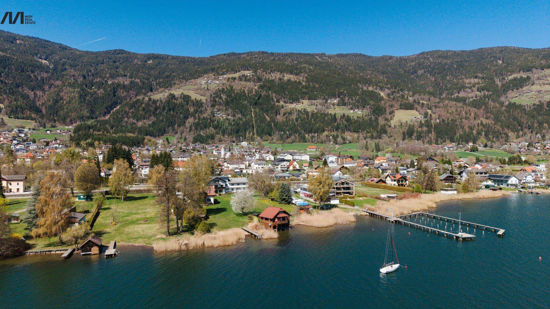 Panoramablick auf die Uferbebauung am See mit Segelboot und bewaldeten Bergen im Hintergrund.