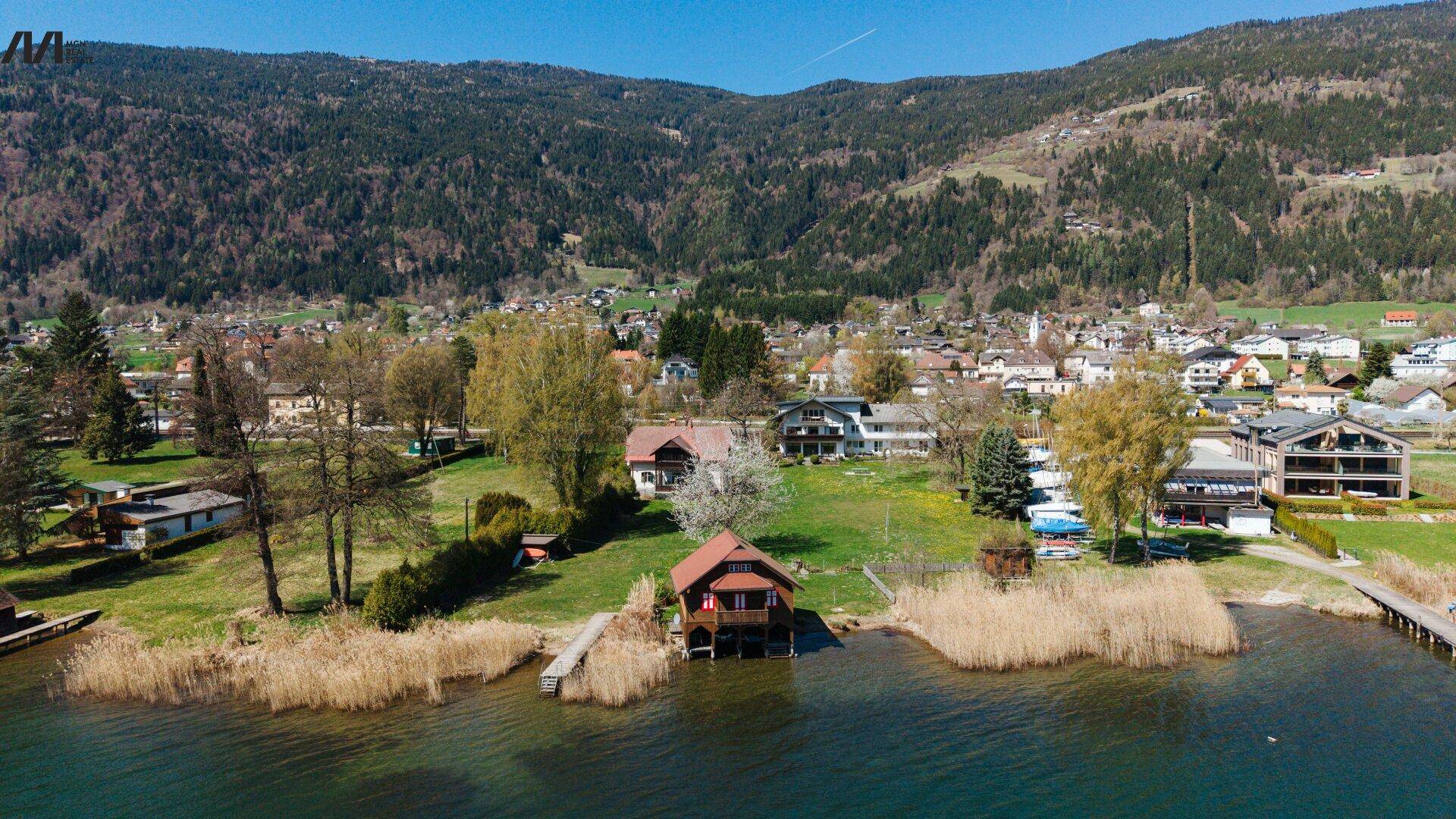Luftaufnahme der malerischen Seeuferlandschaft mit Wohnhäusern, einem Bootshaus und bewaldeten Bergen.