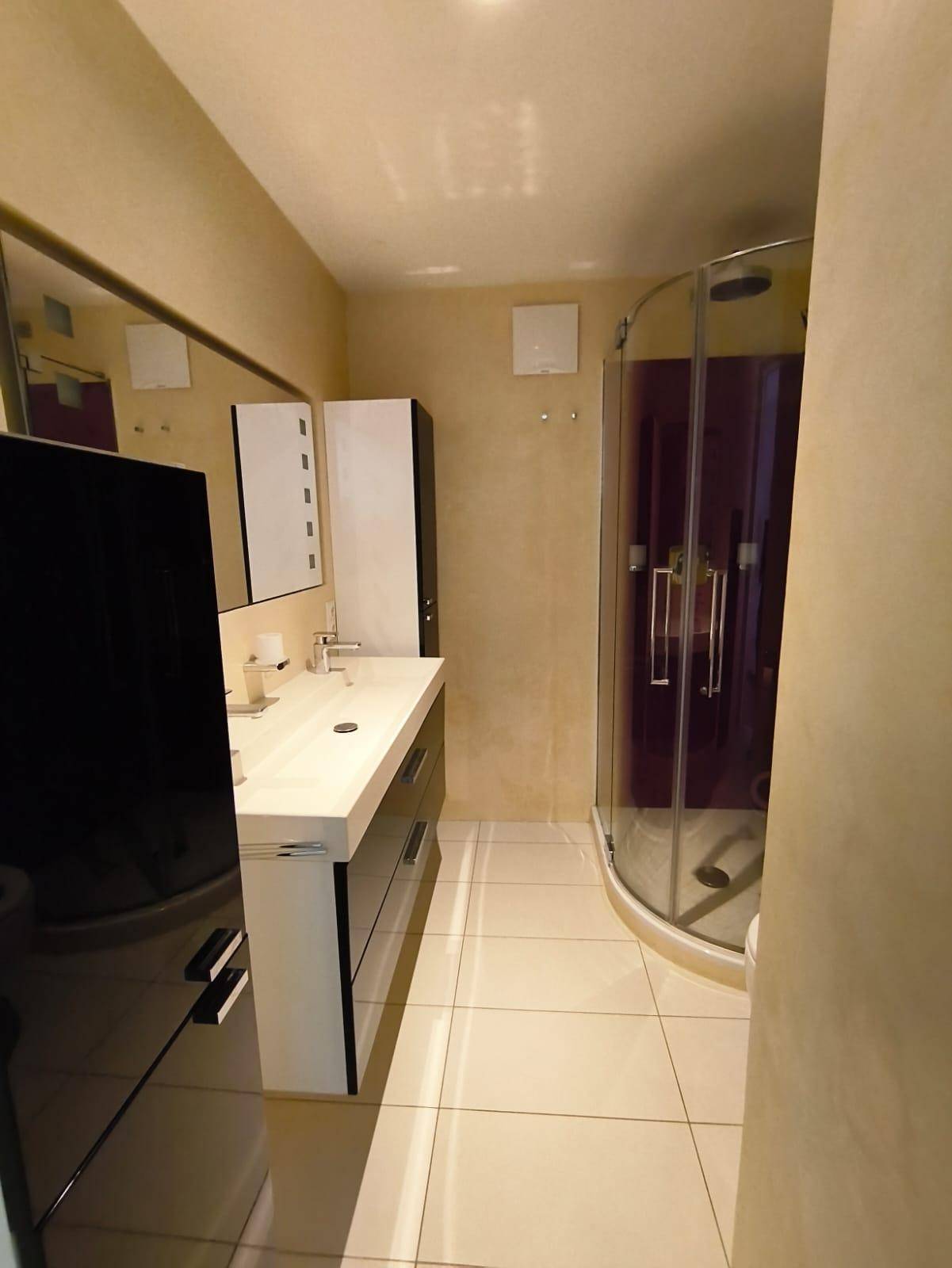 Badezimmer mit Waschtisch, Spiegel und Duschkabine in beigen Farbtönen.