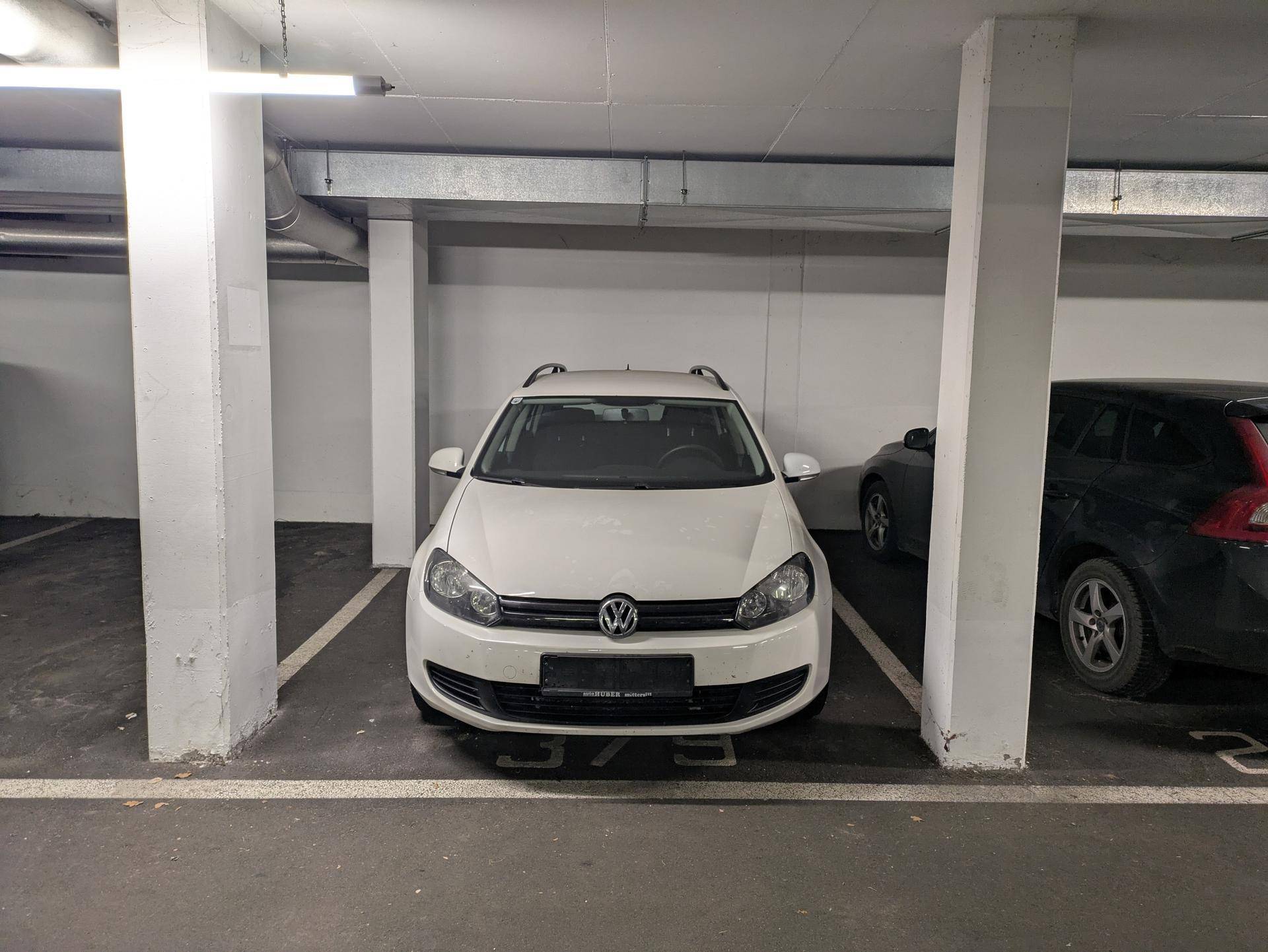 Weißes Auto parkt in einer Tiefgarage mit Betonpfeilern und Beleuchtung.