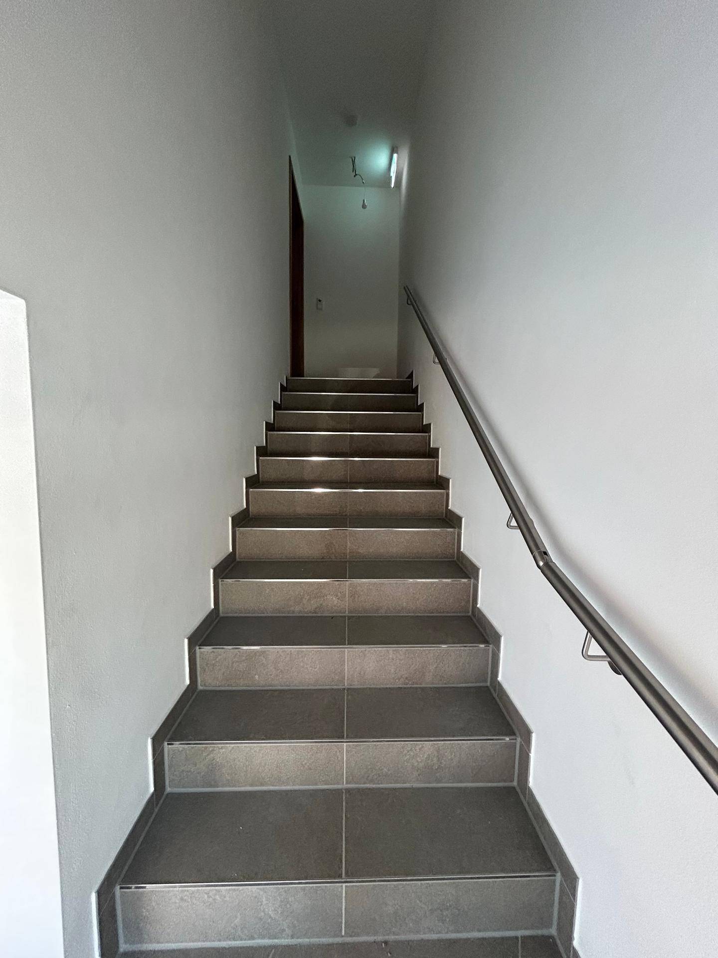 Geräumige Treppe mit grauen Fliesen und einem durchgehenden Edelstahlgeländer, das nach oben führt.