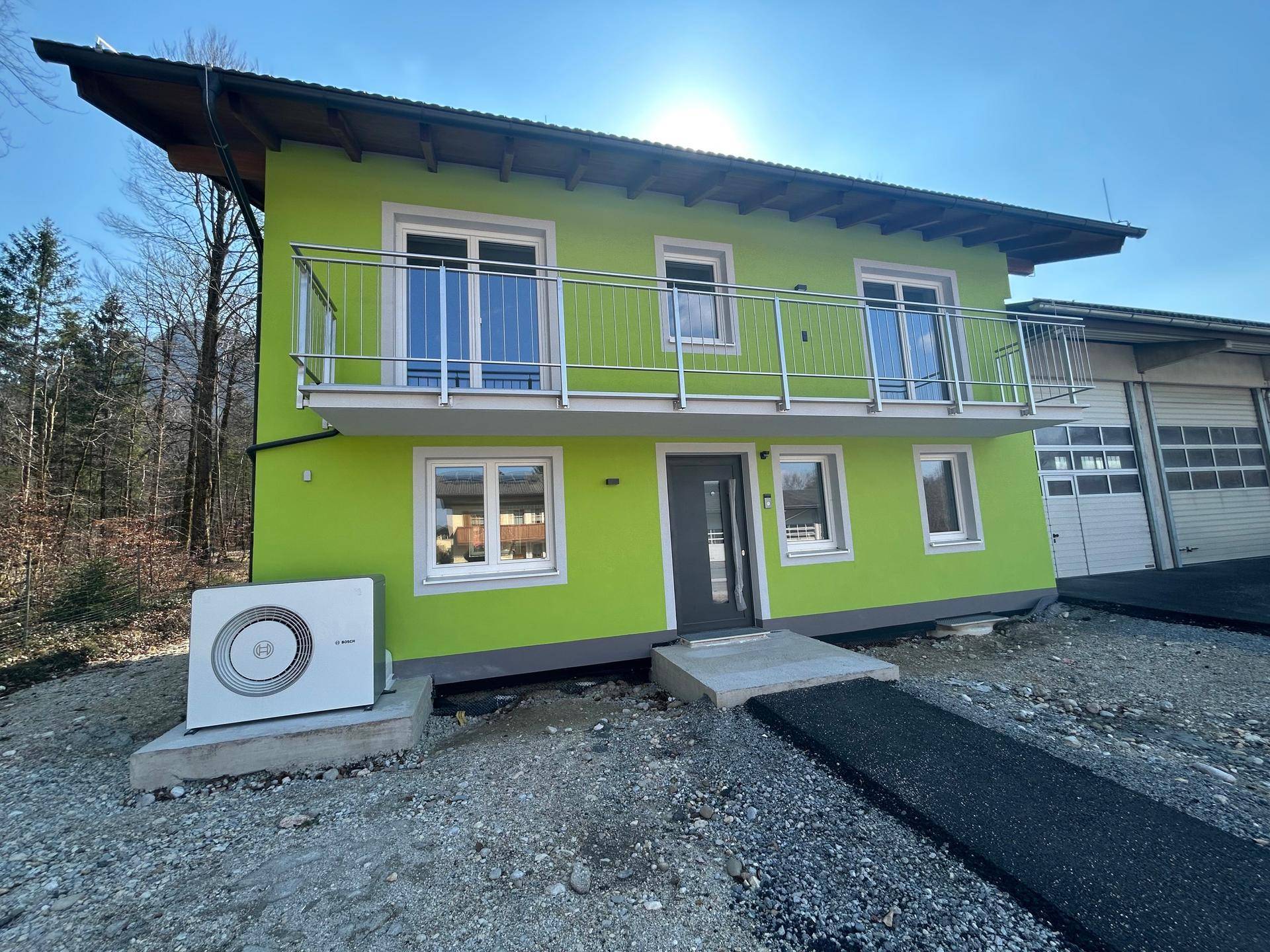 Vorderansicht eines modernen, leuchtend grünen Gebäudes mit Balkon und einer außenliegenden Wärmepumpe.