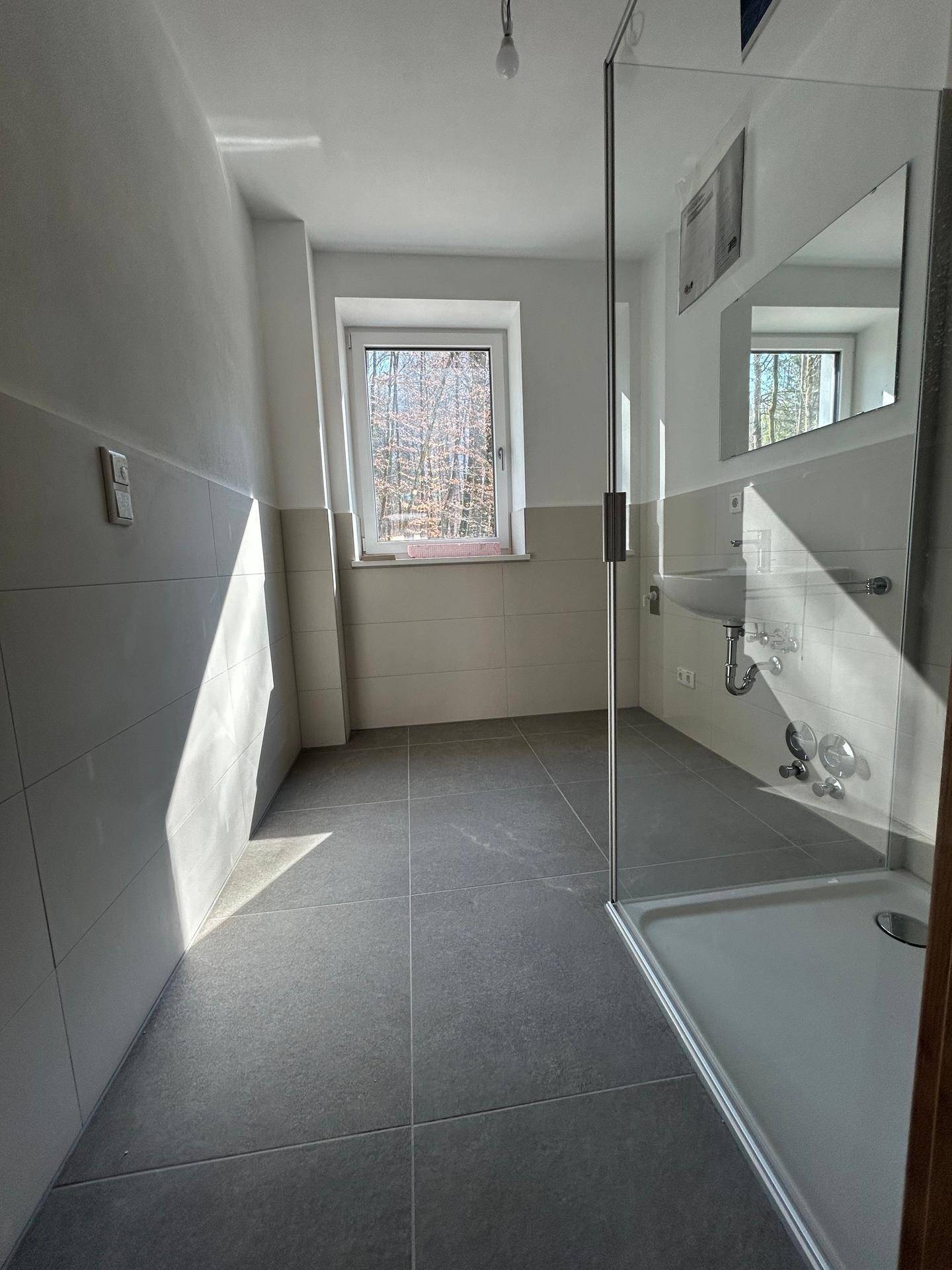 Helles Badezimmer mit Fenster, Duschkabine und grauen Bodenfliesen, die viel Licht reflektieren und den Raum aufhellen.