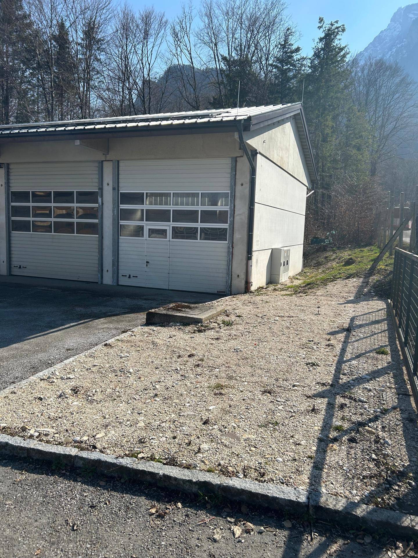 Seitenansicht des Garagengebäudes mit Schotterfläche und angrenzendem Waldgebiet, das eine natürliche Umgebung bietet.