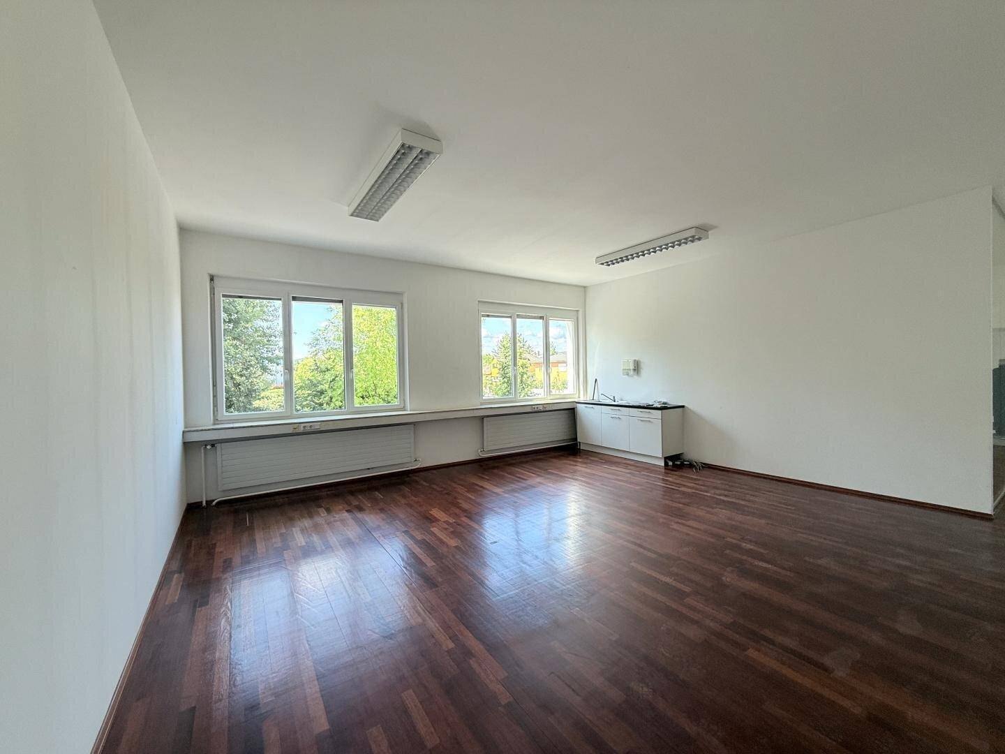 Bürofläche mit dunklem Holzboden, großen Fenstern und einer kleinen Küchenzeile für den täglichen Gebrauch.