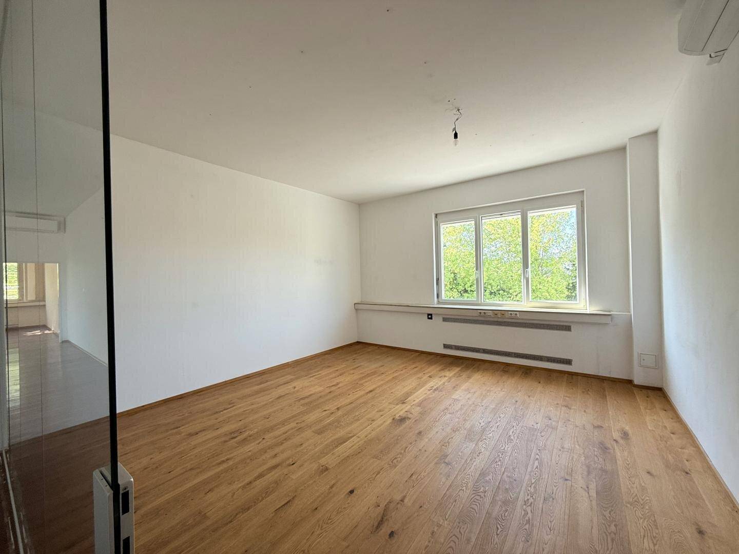 Heller Büroraum mit hellem Holzboden und großen Fenstern, die eine angenehme Arbeitsatmosphäre schaffen.