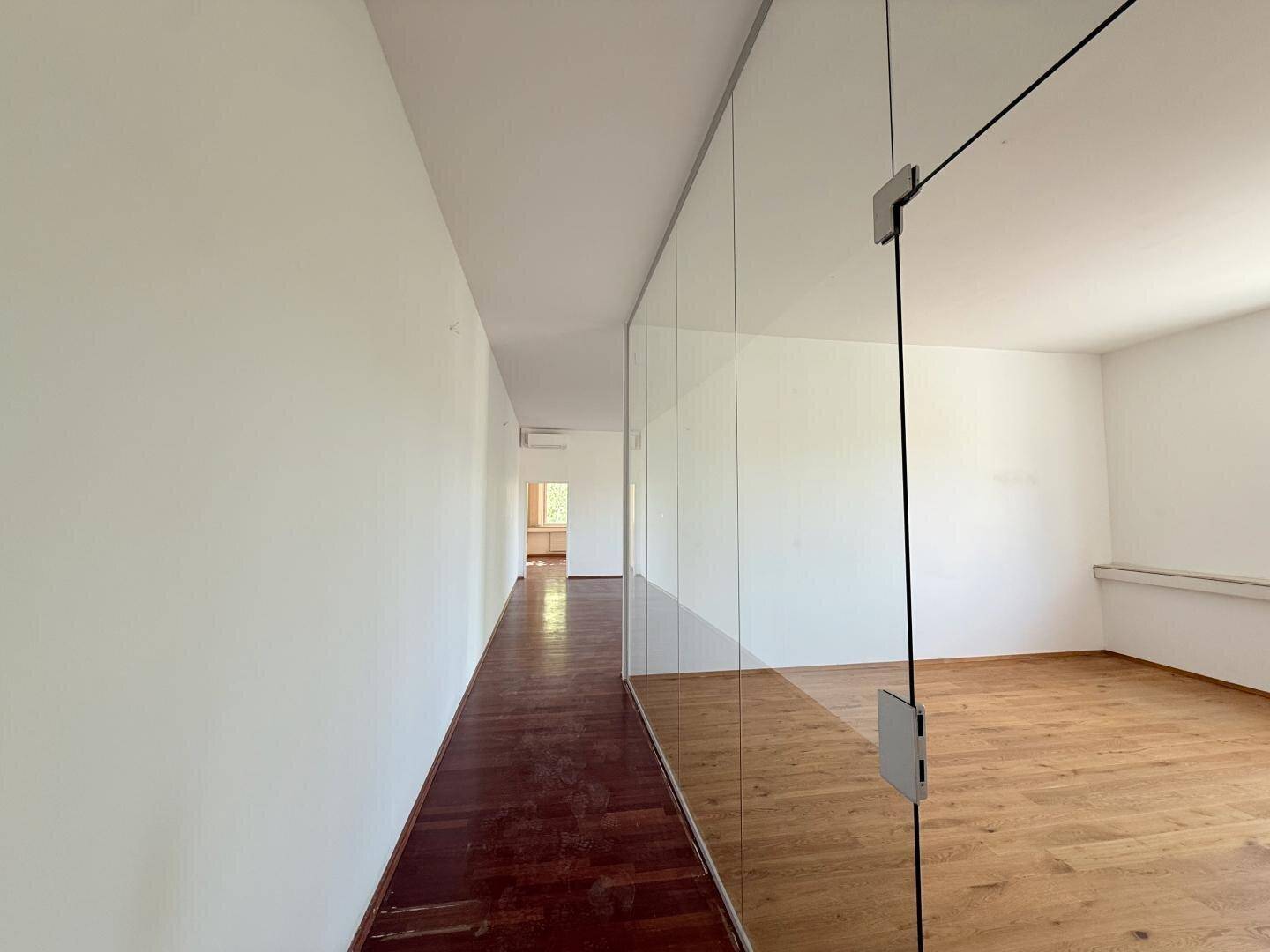 Langer Flur mit dunklem Holzboden und Glaswänden, der zu hellen Büroräumen führt.