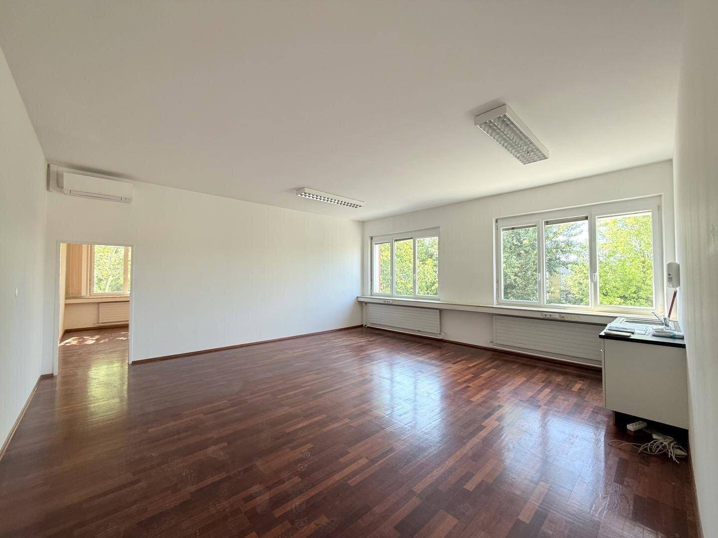 Geräumiges Büro mit dunklem Holzboden und großen Fenstern, die viel Tageslicht hereinlassen.