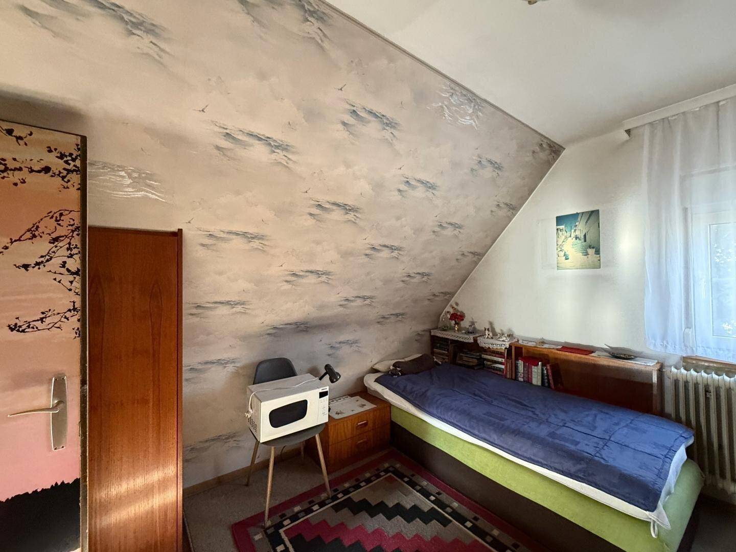 Schlafzimmer mit Dachschräge, gemusterter Tapete und einem Einzelbett, hell beleuchtet.