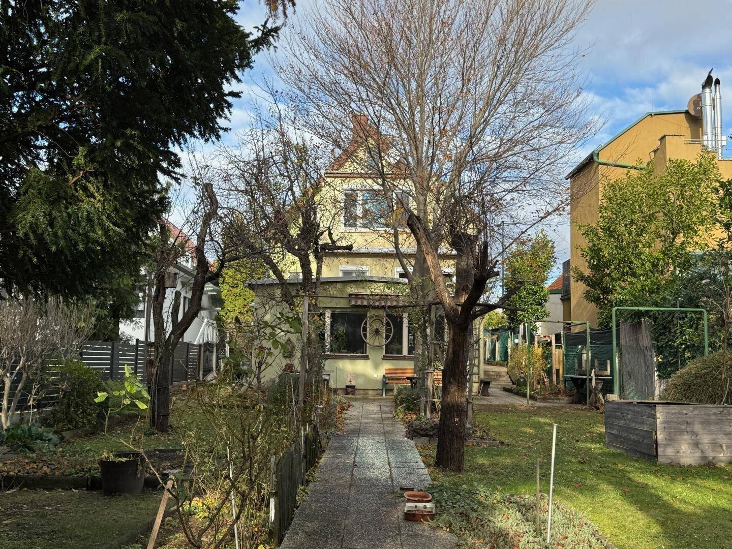 Hausfassade mit Vorgarten und einem gepflasterten Weg, umgeben von Bäumen und Sträuchern.