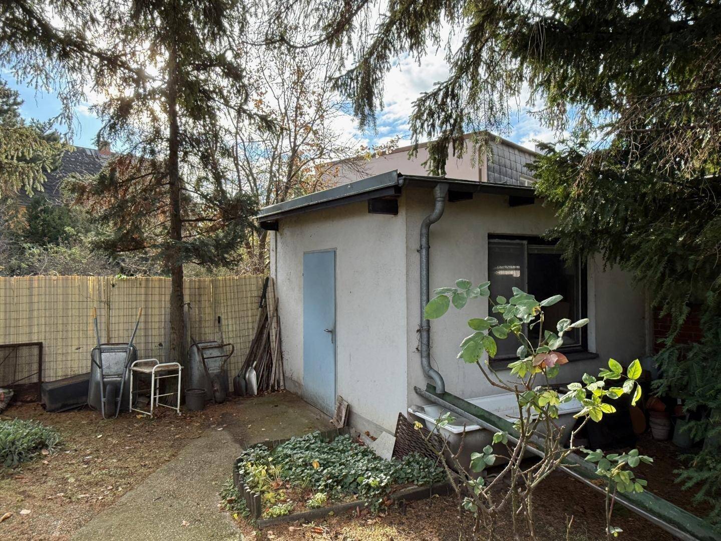 Kleines Gartenhaus oder Schuppen im Gartenbereich, umgeben von Bäumen und Gartengeräten.