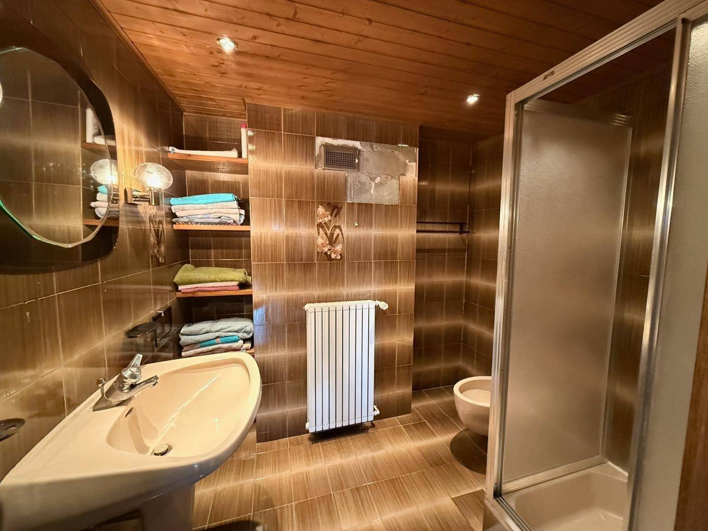Badezimmer mit braunen Fliesen, Dusche, Waschbecken und Holzdecke.