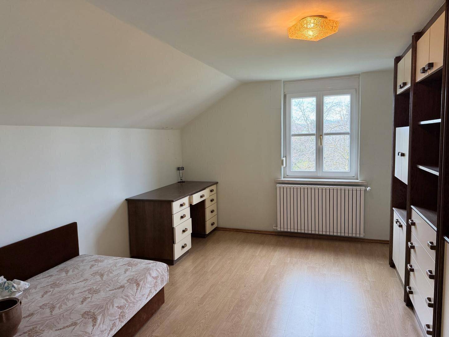 Helles Schlafzimmer mit Dachschräge, Schreibtisch und Fenster mit Ausblick.