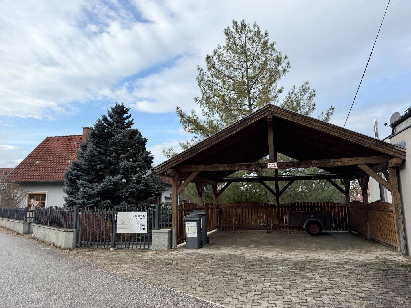 Einfahrt mit Holzcarport und Mülltonnen vor einem traditionellen Haus.