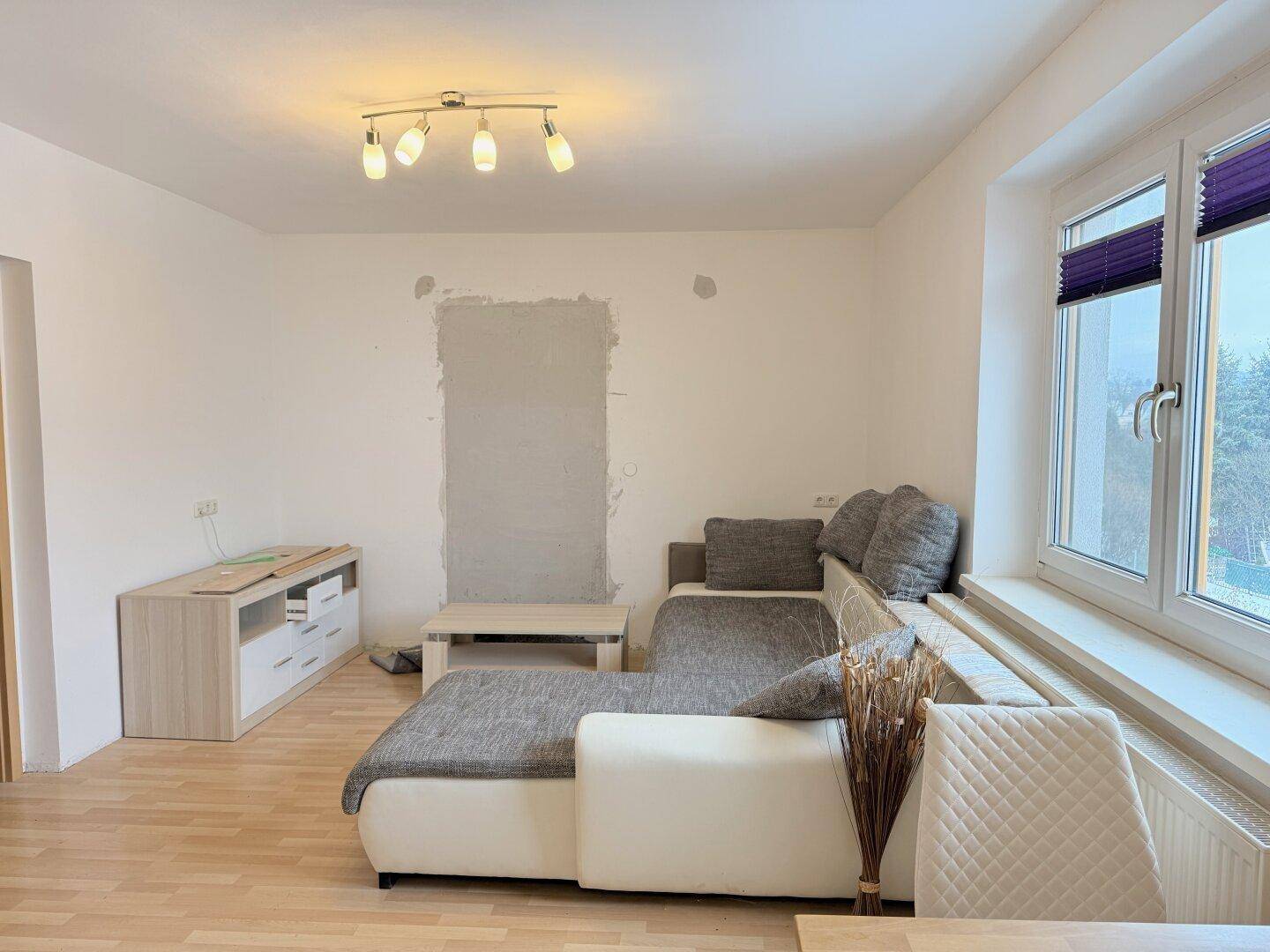 Helles Wohnzimmer mit L-förmigem Sofa, Holzfußboden und großem Fenster, modern eingerichtet.