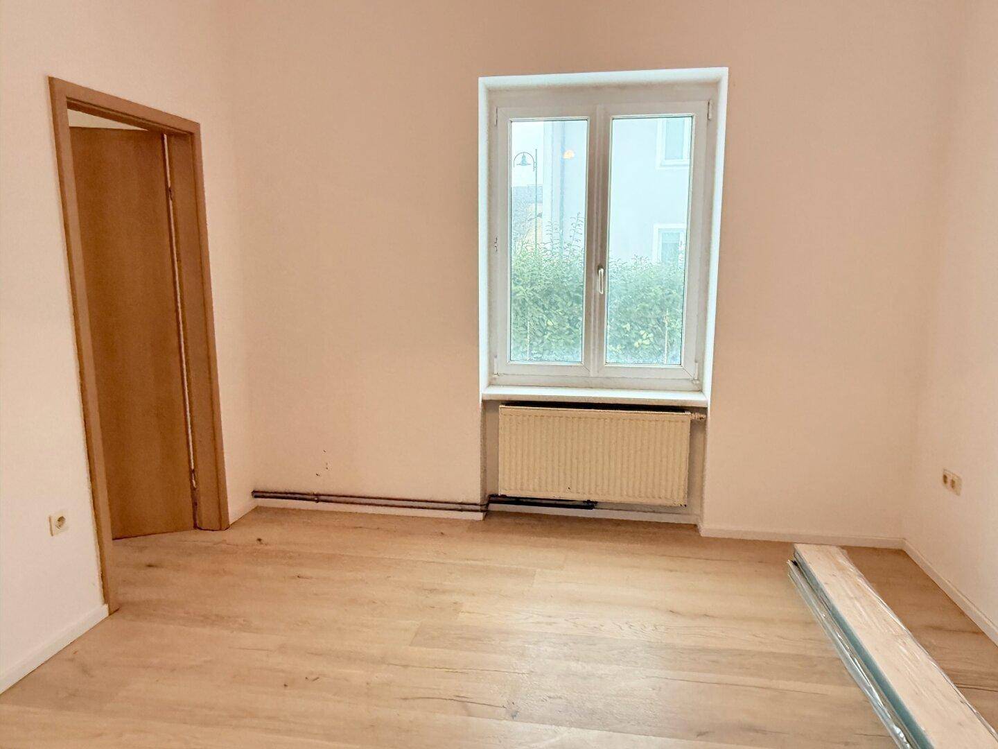 Leeres Zimmer mit neuem Holzfußboden, großem Fenster und Heizkörper, bereit zum Einzug.