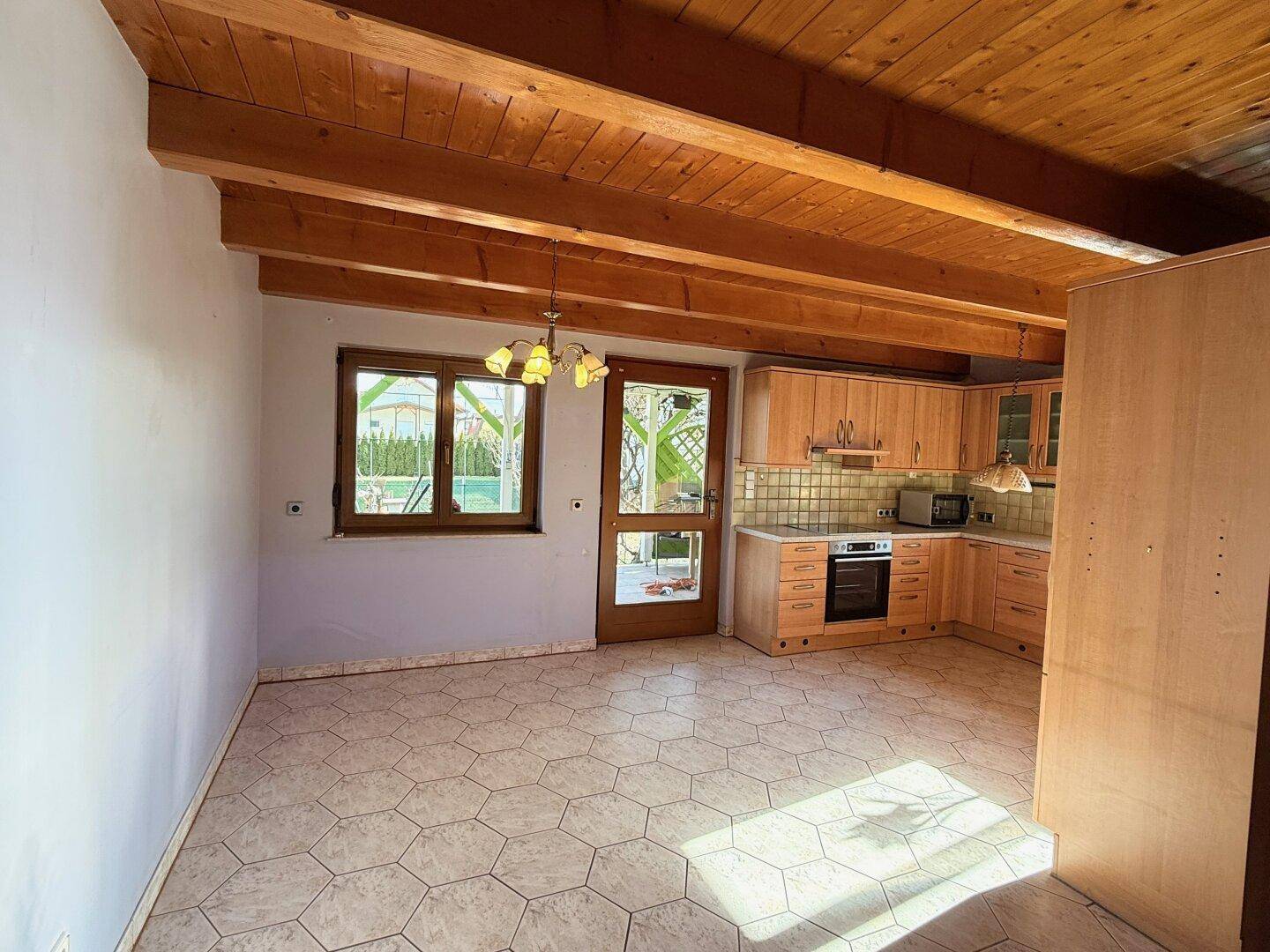 Offener Wohnbereich mit integrierter Holzküche, sichtbaren Holzbalken und Fliesenboden.