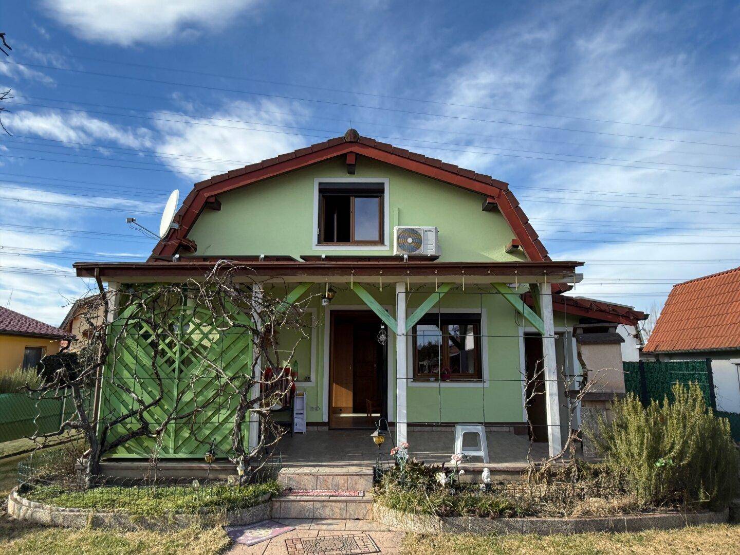 Grünes Einfamilienhaus mit überdachtem Eingangsbereich, Garten und Klimaanlage an der Fassade.