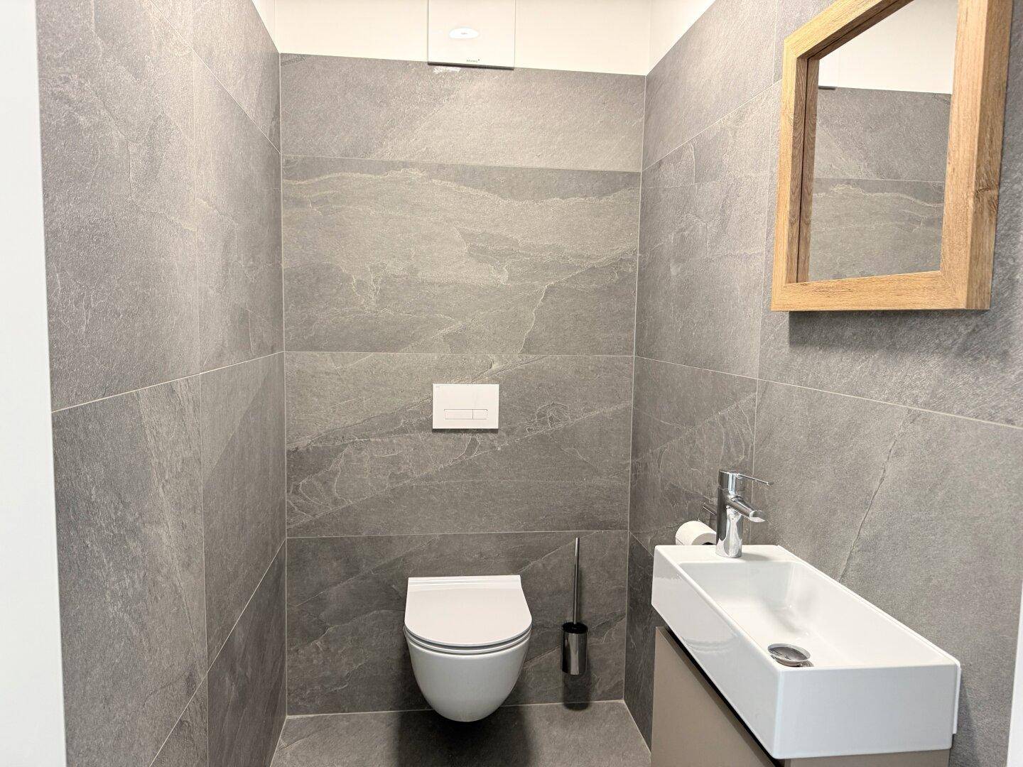Modernes Gäste-WC mit grauen Fliesen, wandhängendem WC und kleinem Waschbecken.