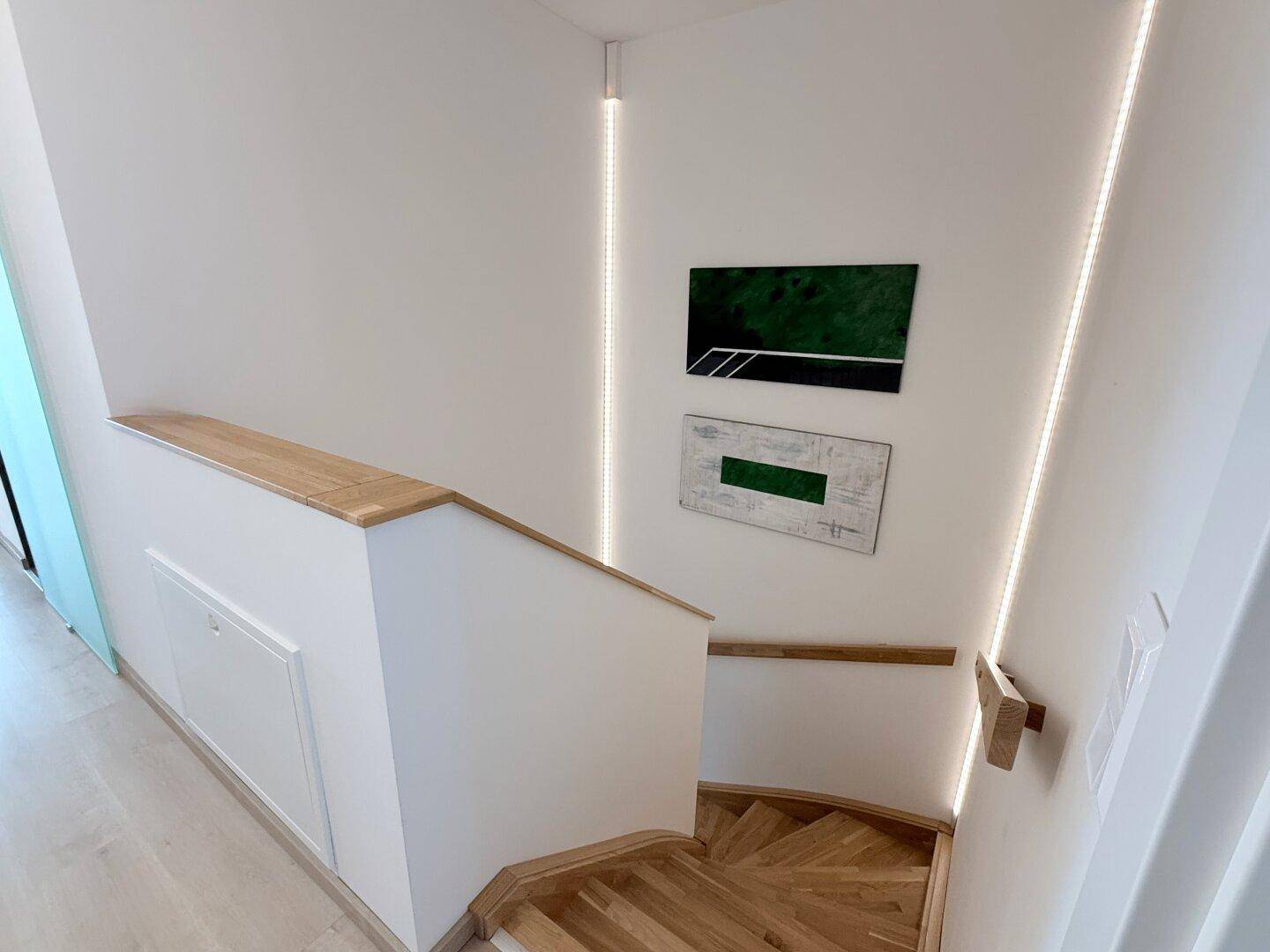 Heller Treppenaufgang mit Holzstufen, weißem Geländer und indirekter Beleuchtung.