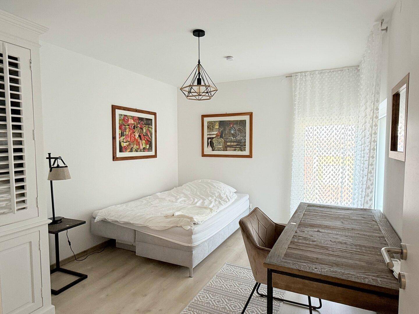 Helles Schlafzimmer mit Einzelbett, Schreibtisch und Fenster mit Vorhängen.