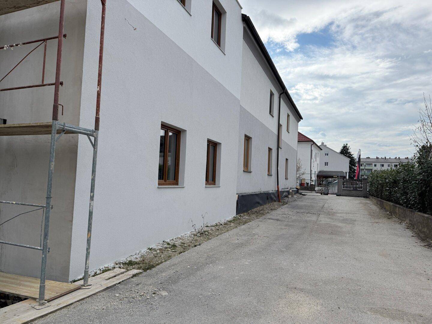 Lange Seitenansicht eines Neubaus mit zweifarbiger Fassade und Zufahrt unter bewölktem Himmel.