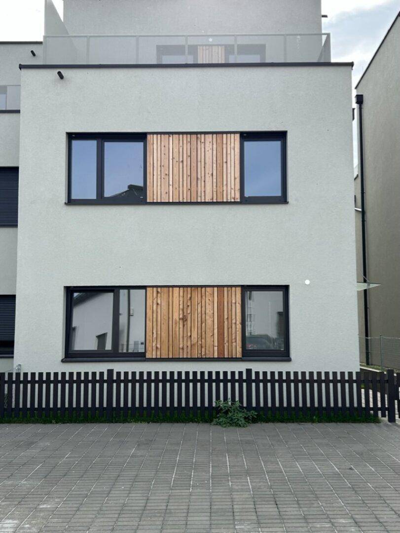 Moderne Gebäudefassade mit hellgrauen Wänden, schwarzen Fensterrahmen und Holzverkleidungen.