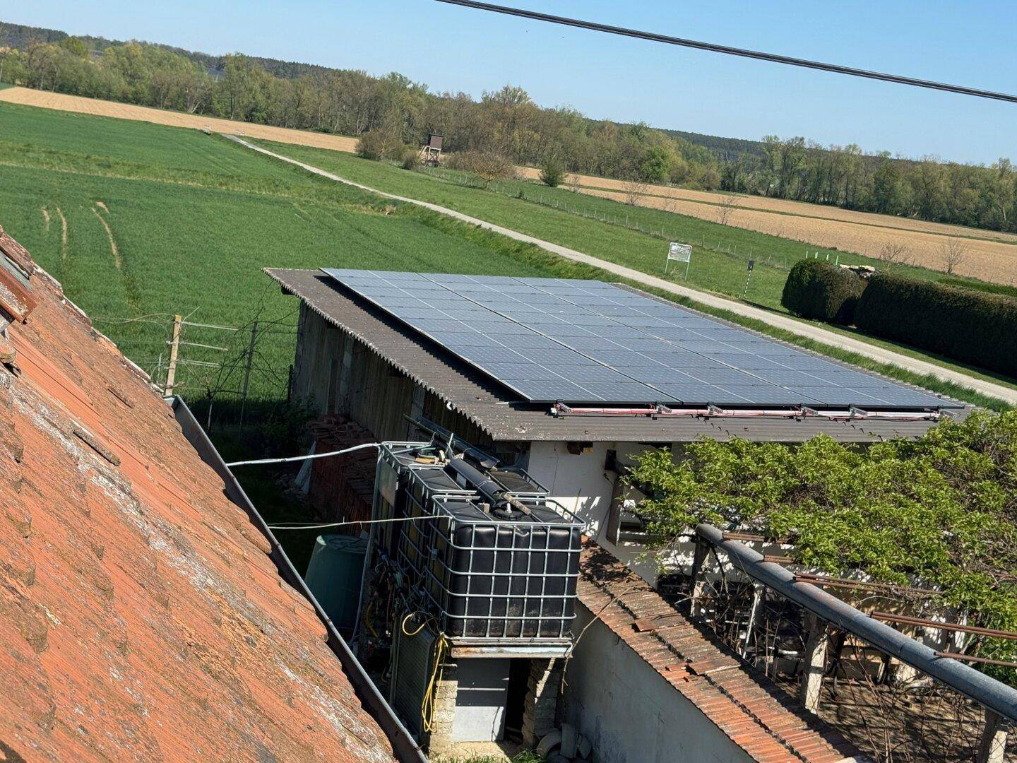 Dachansicht eines Gebäudes mit Photovoltaikanlage und Wassertank, umgeben von Feldern und Natur.