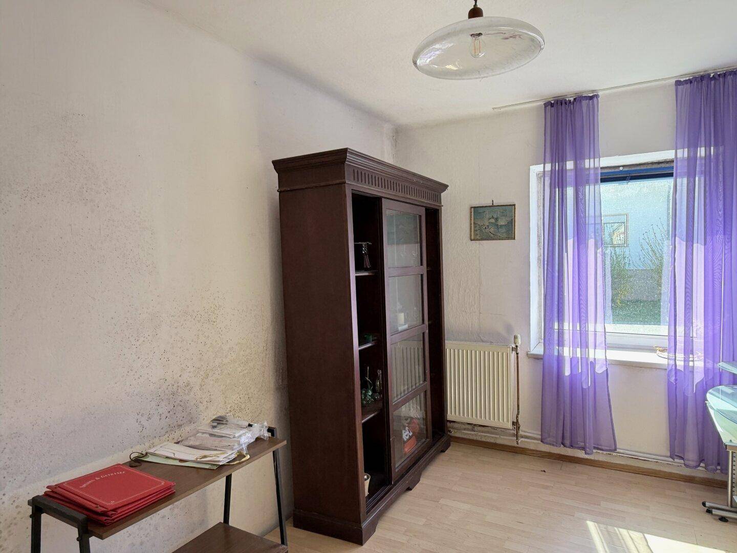 Helles Zimmer mit lila Vorhängen, einem Holzschrank und einem Fenster mit Tageslicht.
