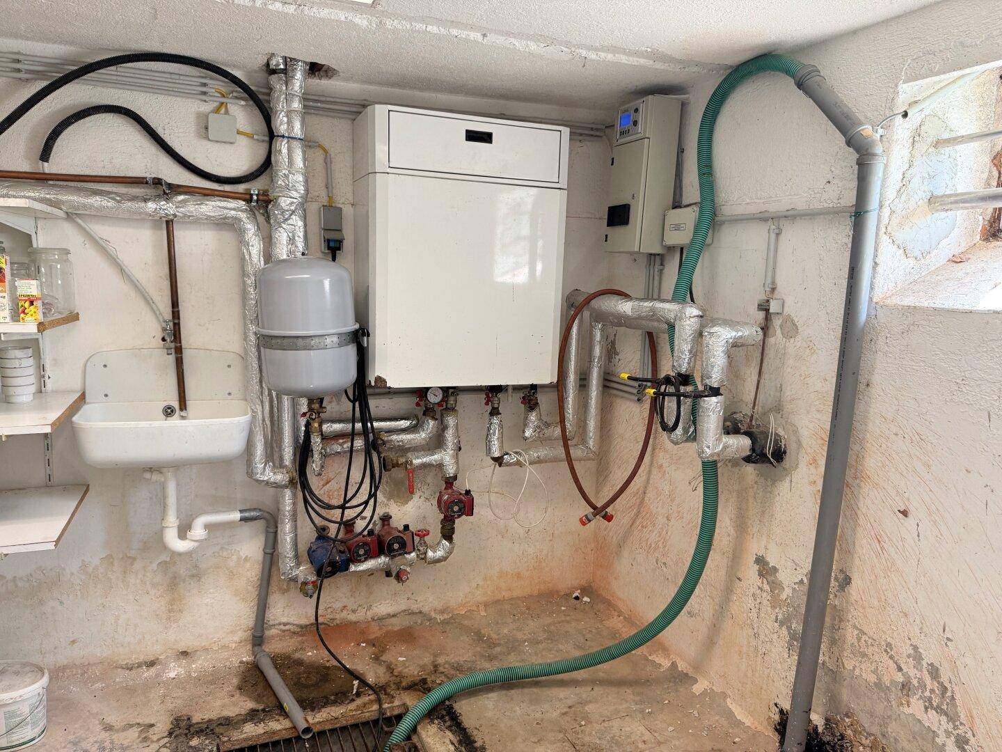 Heizungsraum im Keller mit Warmwasserbereiter, Rohrleitungen und einem kleinen Waschbecken.