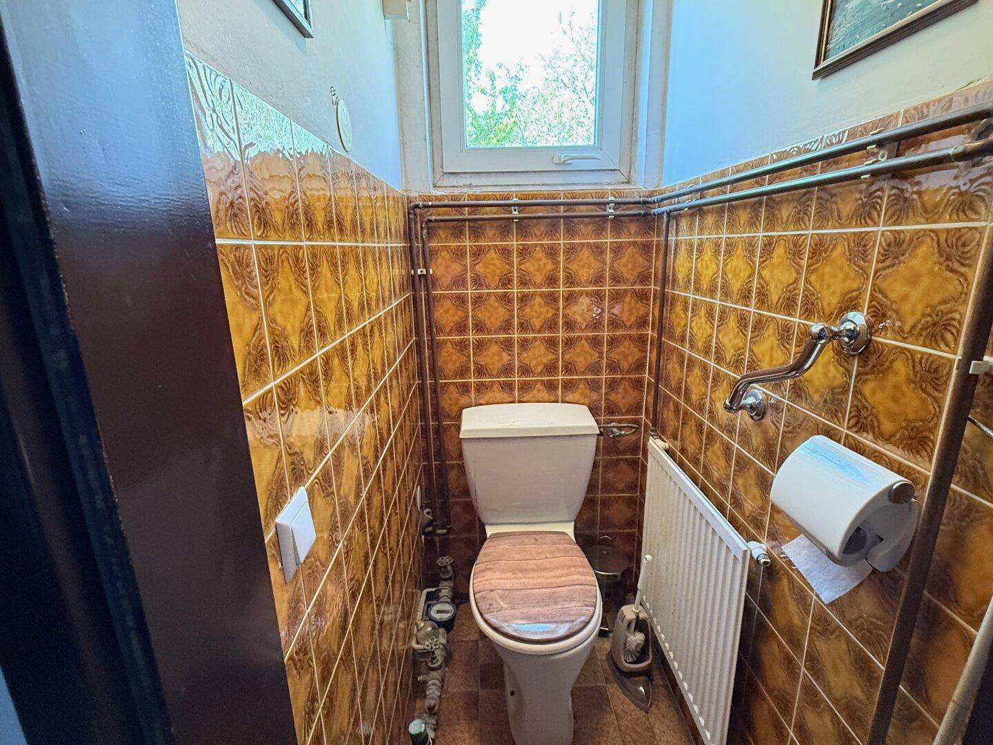 Kleines WC mit Toilette, Heizkörper und braunen Fliesen an den Wänden und auf dem Boden.