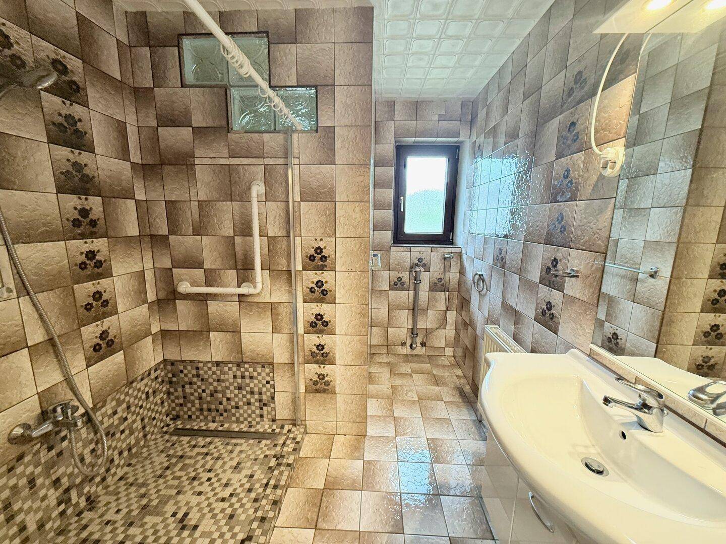 Badezimmer mit barrierefreier Dusche, braunen Fliesen, Haltegriffen und Fenster für Tageslicht.
