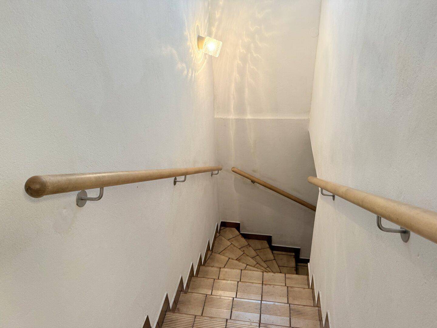 Treppenhaus mit hellem Holzgeländer und gefliesten Stufen, gut beleuchtet.