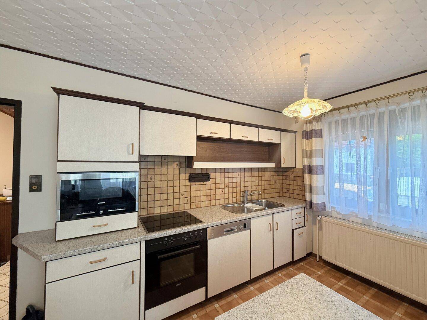 Traditionelle Einbauküche mit hellen Fronten, Induktionskochfeld, Backofen und Fenster mit Vorhängen.