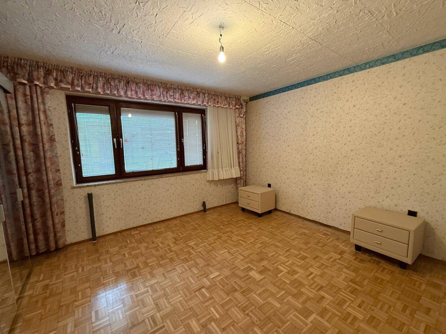 Schlafzimmer mit Parkettboden, gemusterter Tapete und großem Fenster mit Jalousien.