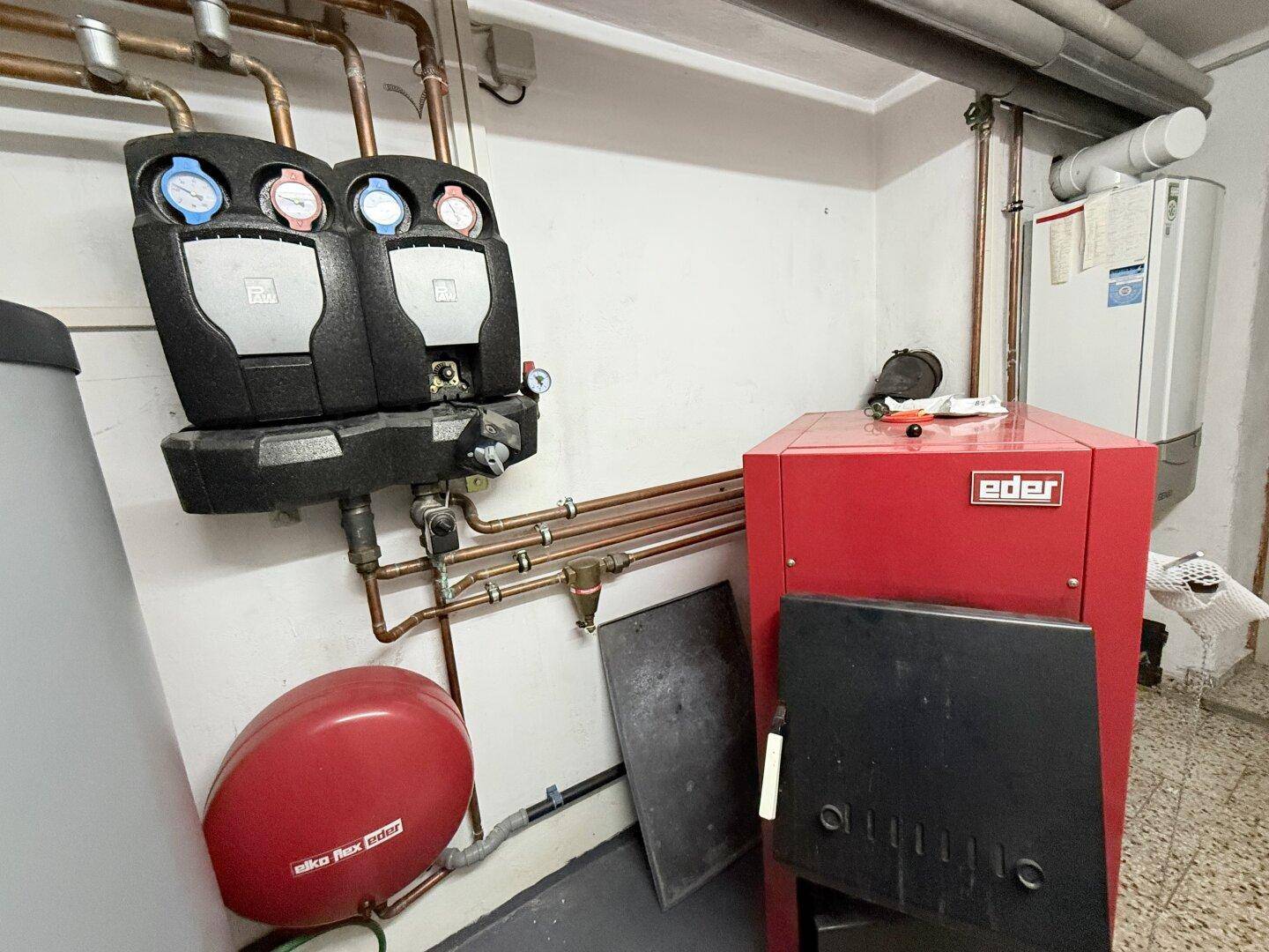 Heizungsraum im Keller mit rotem Heizkessel, Warmwasserspeicher und sichtbaren Rohrleitungen.