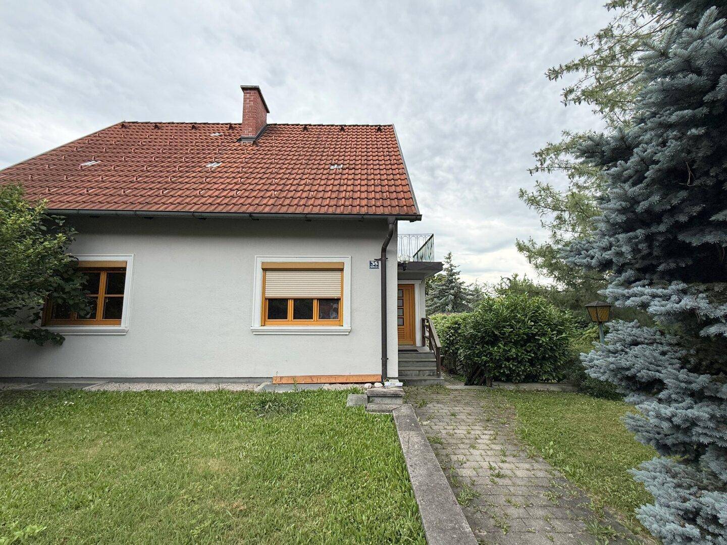 Seitenansicht des Hauses mit einem gepflasterten Weg, Rasenfläche und einem großen Nadelbaum.