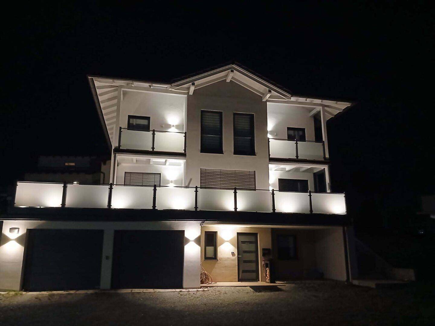 Modernes Einfamilienhaus bei Nacht, beleuchtet mit mehreren Balkonen und integrierter Doppelgarage.