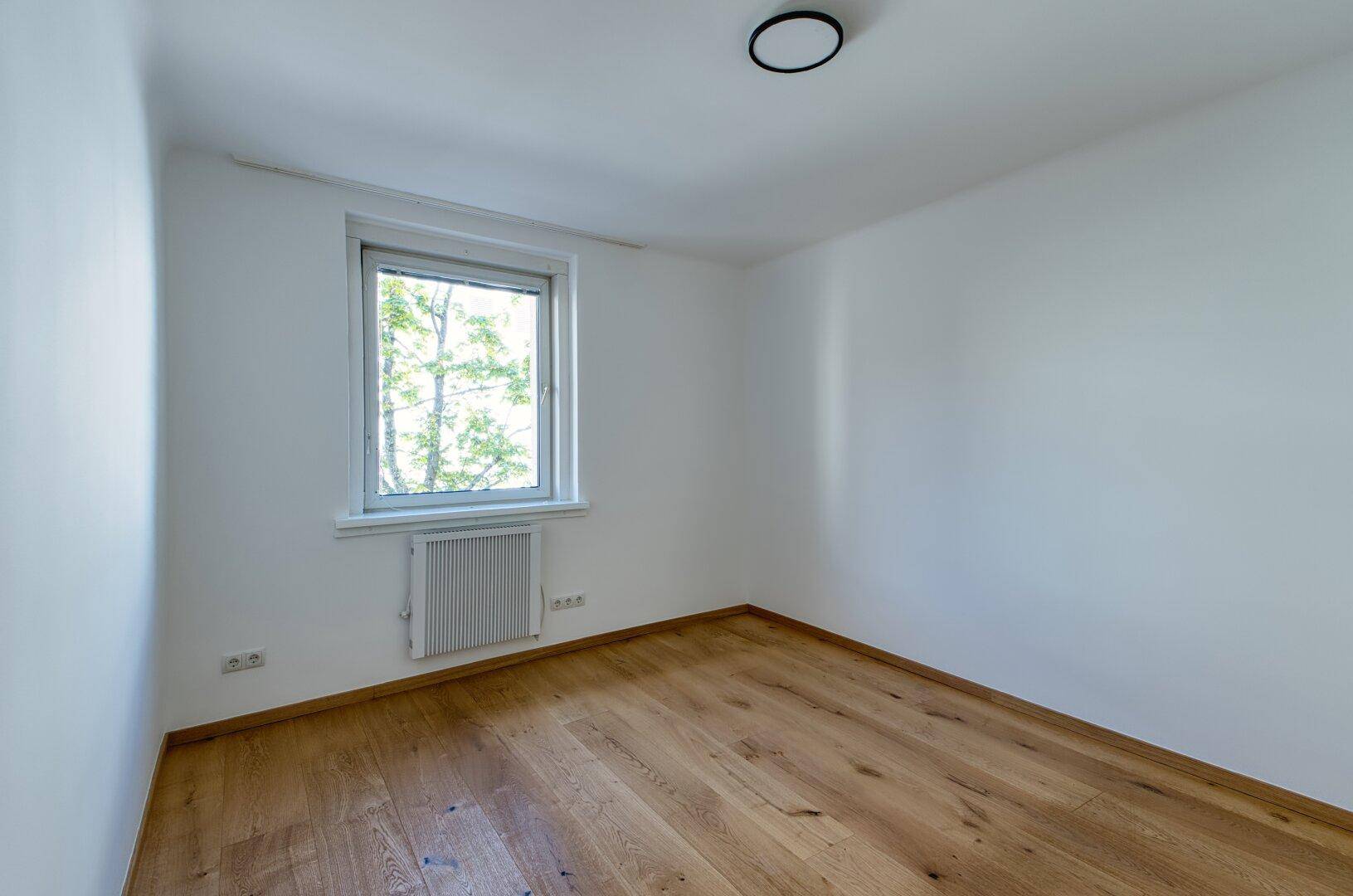 Helles Zimmer mit Holzboden und Fenster, das einen angenehmen Blick ins Grüne bietet.