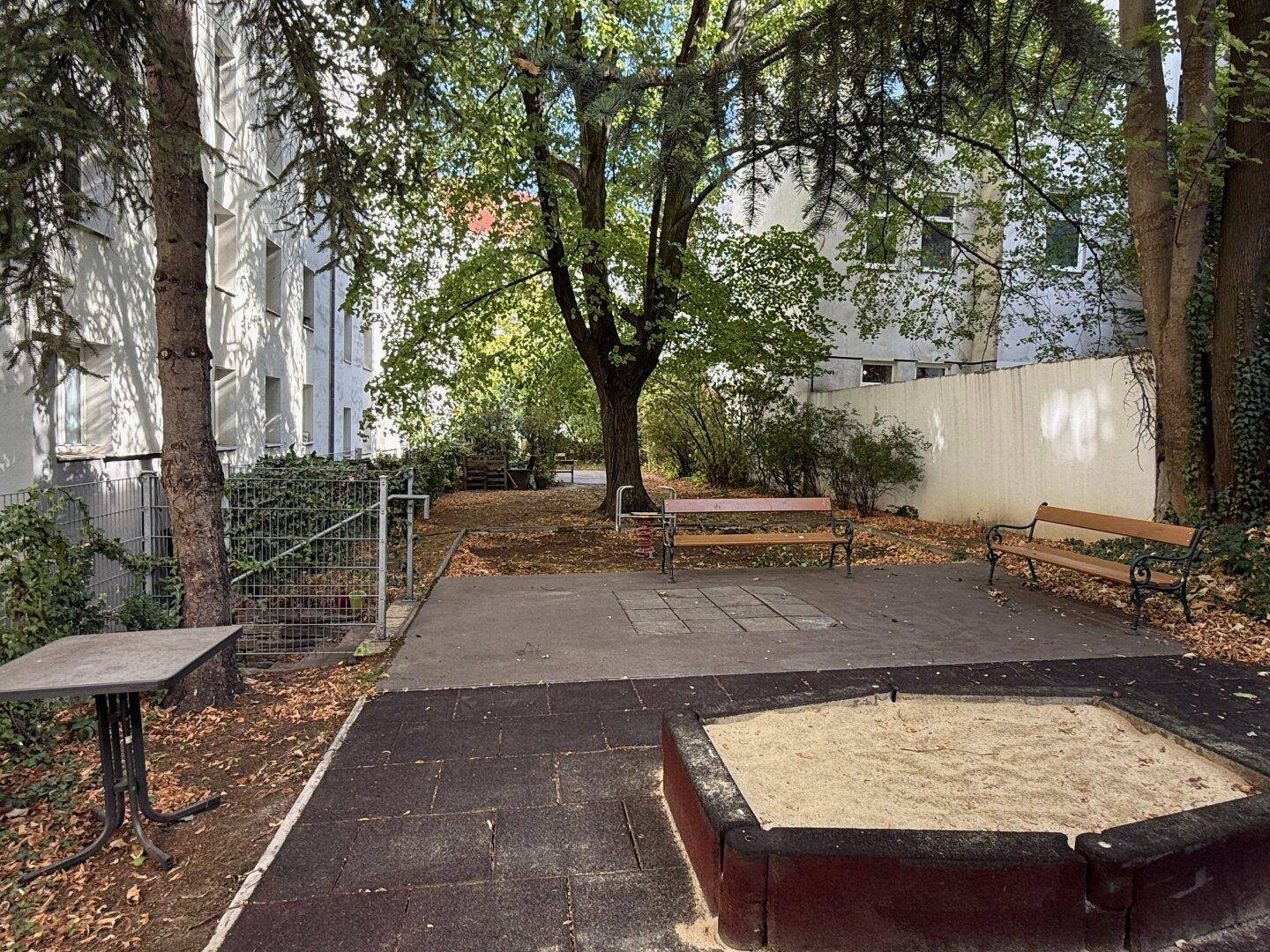 Gemeinschaftlicher Gartenbereich mit Sandkasten und Sitzbänken unter schattenspendenden Bäumen.