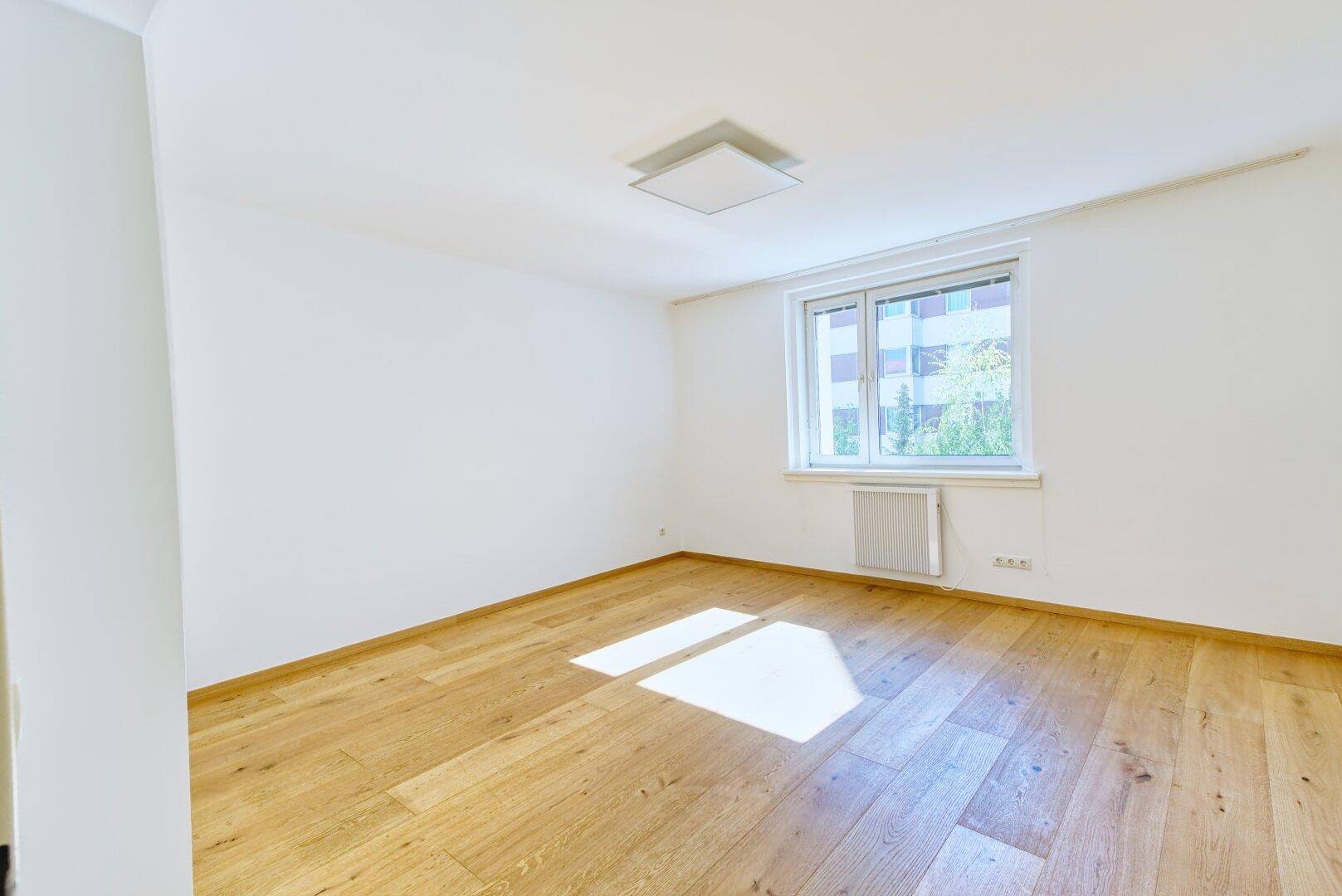 Helles, leeres Zimmer mit Holzboden und großem Fenster, das viel Tageslicht hereinlässt.