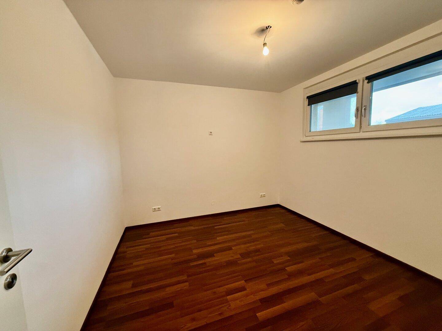 Helles Schlafzimmer mit dunklem Parkettboden und Fenster mit Rollladen.