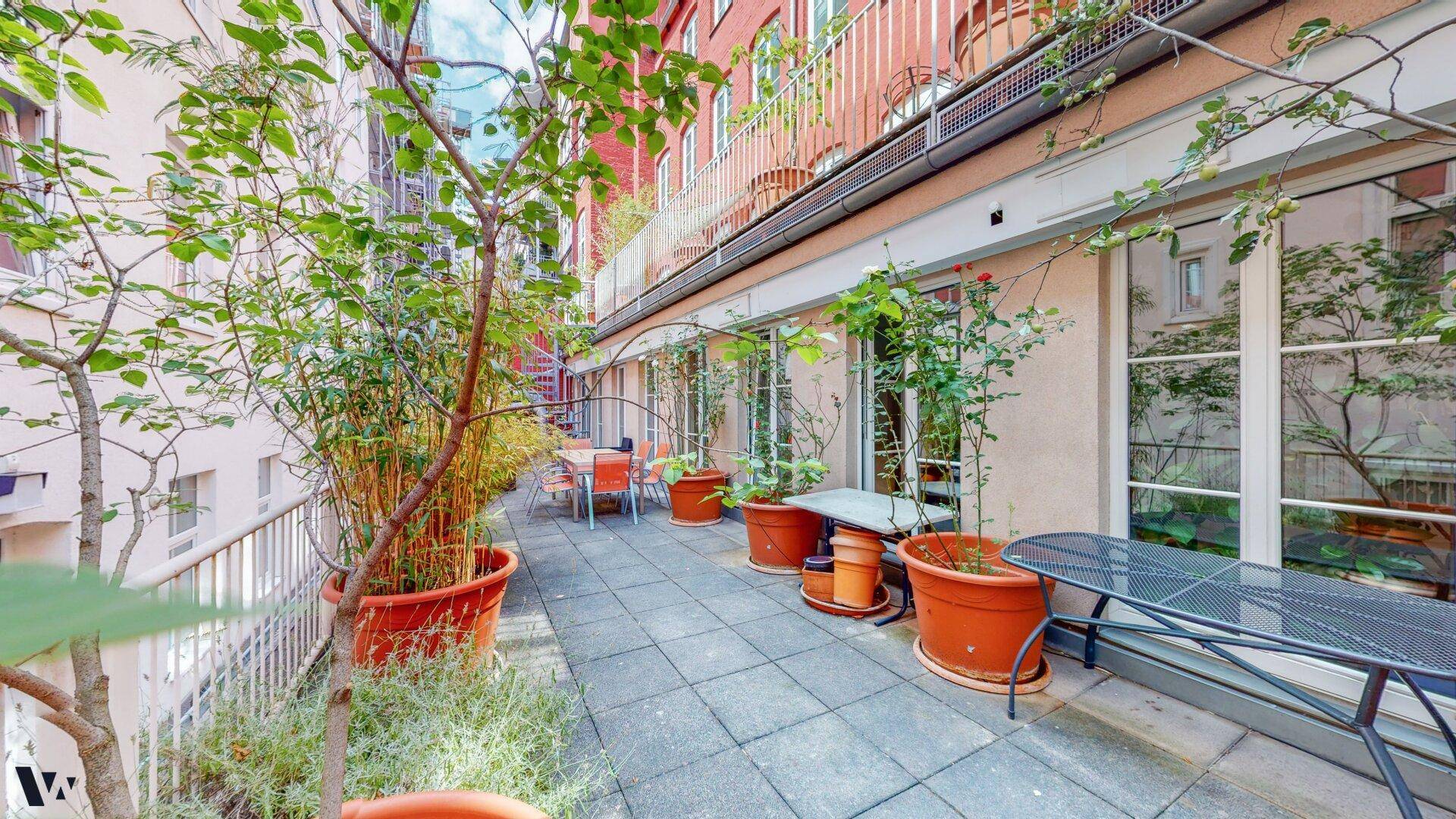 Geräumige Terrasse mit Fliesenboden, Pflanzen in Töpfen und Gartenmöbeln, umgeben von Gebäuden.