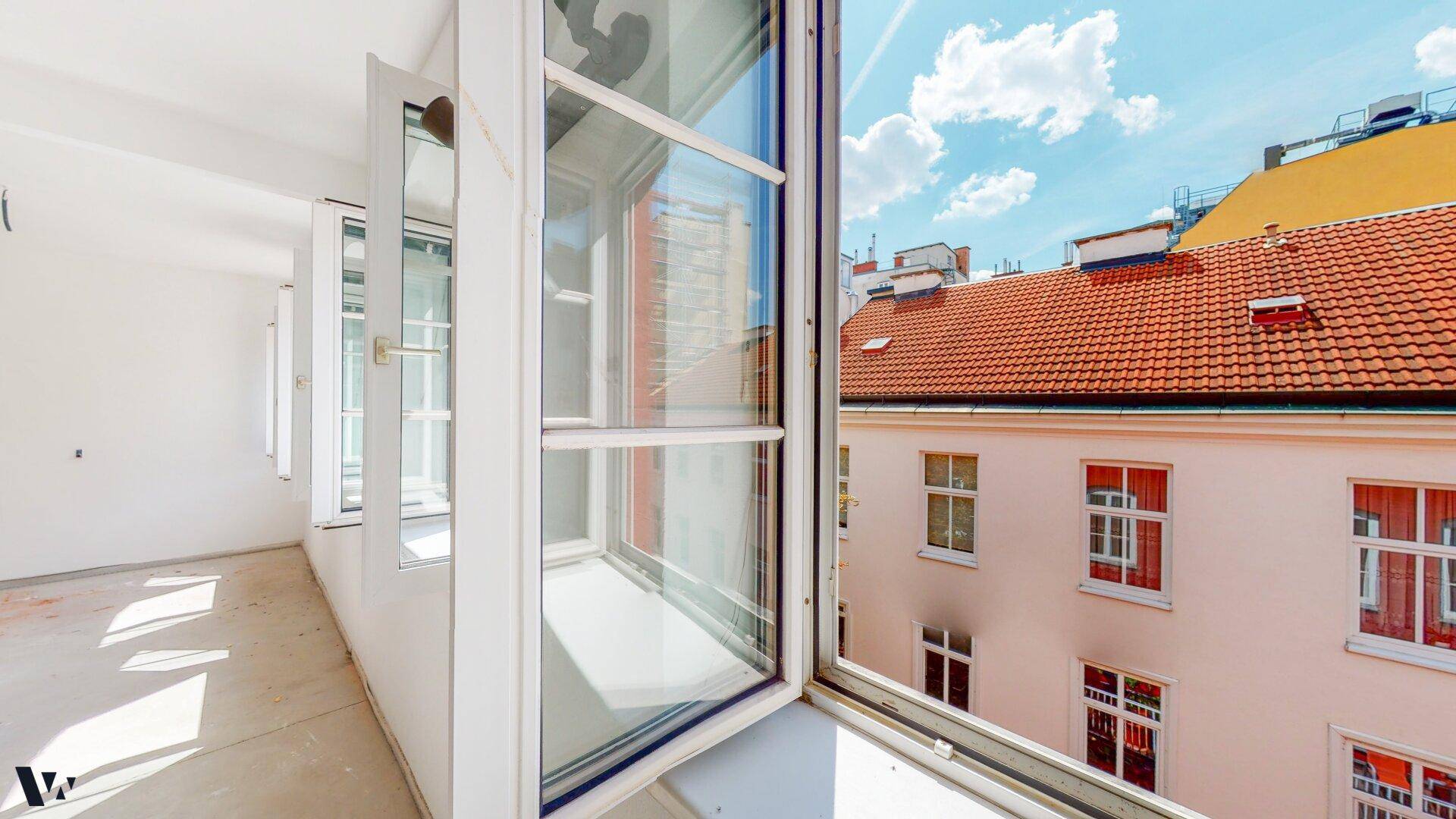 Blick aus einem geöffneten Fenster auf die Dächer der Nachbargebäude und den blauen Himmel.