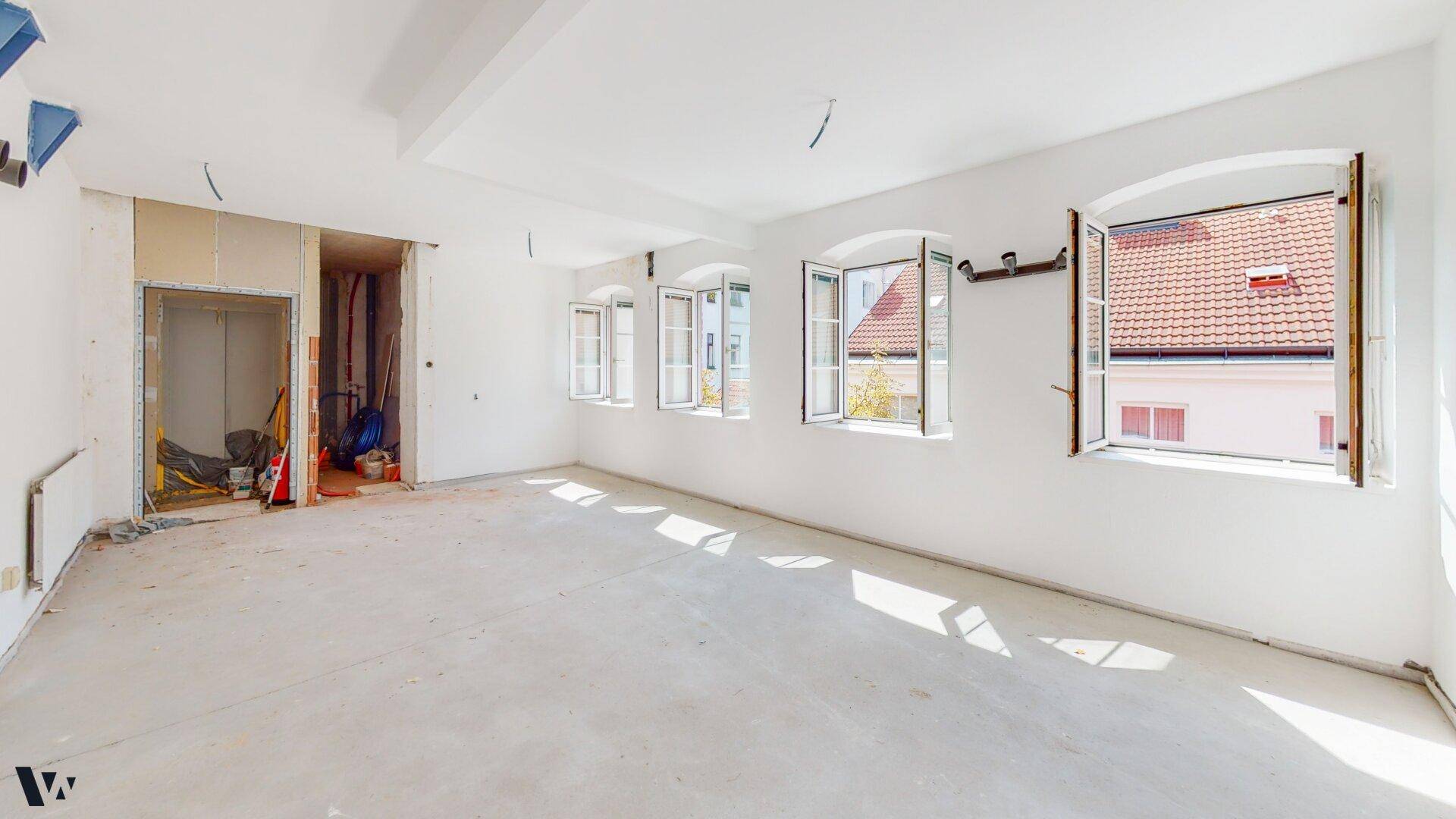 Großer Raum im Rohbau mit Betonboden und vielen Fenstern, ideal für individuelle Gestaltung.