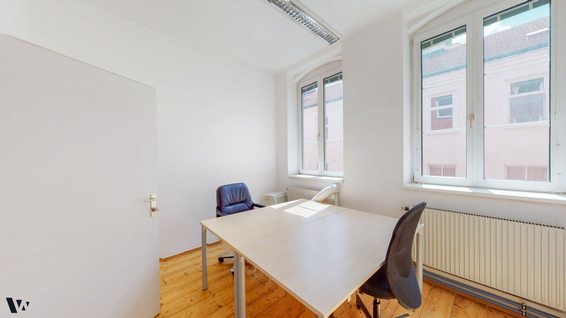 Helles Einzelbüro mit Holzdielenboden, einem Schreibtisch und zwei Fenstern.