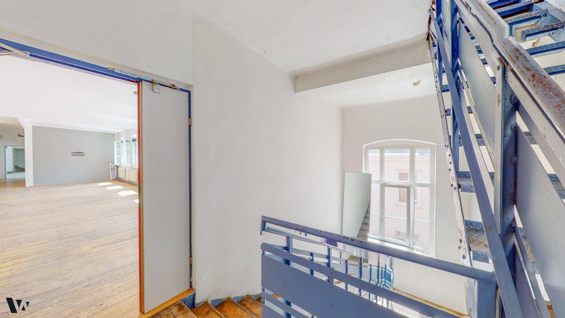 Treppenhaus mit blauen Metallgeländern und Holzdielen, das zu den oberen Etagen führt.