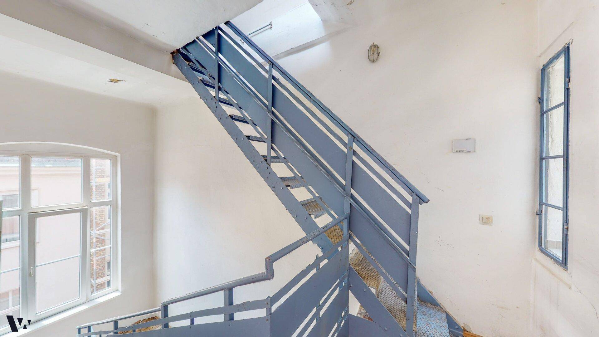 Industriell anmutendes Treppenhaus mit blauen Metallgeländern und großen Fenstern.