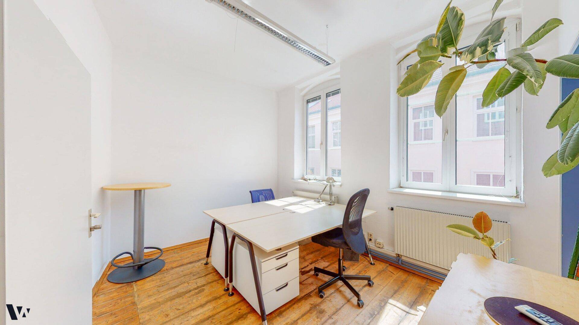 Büro mit Holzdielenboden, zwei Arbeitsplätzen und einem Stehtisch in der Ecke.